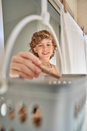 Foto de Niño feliz con el pelo rubio rizado sonriendo y tomando pinzas de ropa para secar la ropa blanca mientras hace las tareas domésticas en casa - Imagen libre de derechos