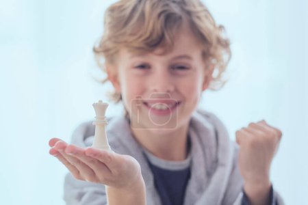 Téléchargez les photos : Joyeux garçon préadolescent démontrant pièce d'échecs de la reine et tenant le poing vers le haut se réjouissant de la victoire - en image libre de droit