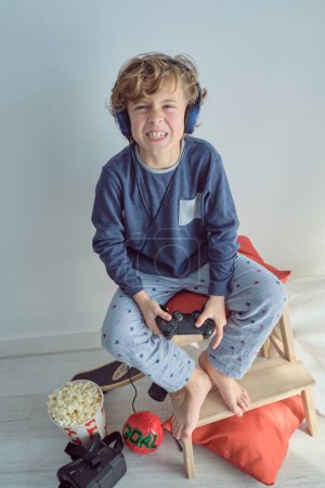 Full length from above of adorable barefoot blond haired kid in headphones and pajama baring teeth while playing video game with joystick against white wall and looking at camera