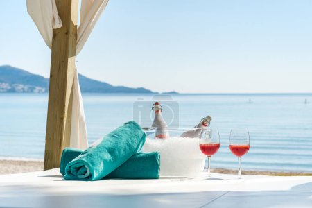 Foto de Botellas y par de gafas de vino con vino de rosas frías colocadas con toallas de playa enrolladas en la cama con dosel para una velada romántica en la orilla del mar - Imagen libre de derechos