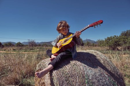 Foto de Cuerpo completo de chico descalzo tocando guitarra acústica y practicando acordes de melodía en tiempo libre sobre pacas de heno en ambiente rural - Imagen libre de derechos