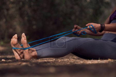 Photo for Side view of crop unrecognizable barefooted female athlete in sportswear sitting on ground and stretching legs with resistance band during outdoor training in forest on sunny day - Royalty Free Image