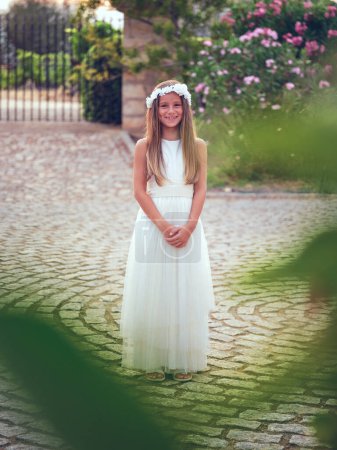 Photo for Full body of cheerful kid wearing white dress and flower wreath standing on pavement near green trees in garden while smiling at camera at sunset - Royalty Free Image