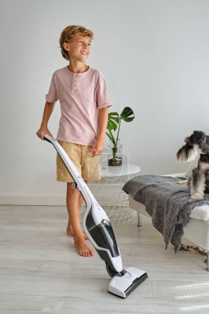 Photo for Full body barefoot boy smiling and looking away while using vacuum cleaner to tidy floor near chair with Schnauzer dog in light living room at home - Royalty Free Image