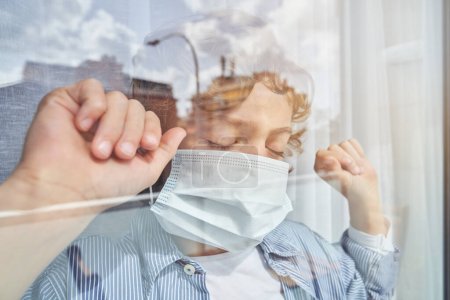 Photo for Through glass view of boy with curly blond hair in casual clothes and medical mask standing near window with closed eyes and clenched fists during COVID pandemic - Royalty Free Image