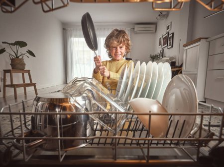 Photo for Content boy with frying pan in hands standing near opened dishwasher with various clean dishware during household routine in kitchen - Royalty Free Image