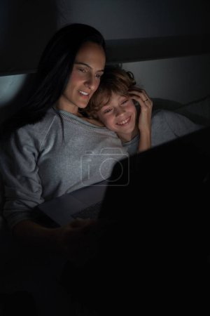 Foto de Sonriente chico con la madre navegando por Internet en netbook mientras hacen la tarea juntos en la cama en dormitorio oscuro tarde en la noche - Imagen libre de derechos
