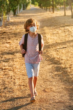 Foto de Cuerpo completo de niño con máscara protectora y caminando por sendero herboso durante el brote de coronavirus - Imagen libre de derechos