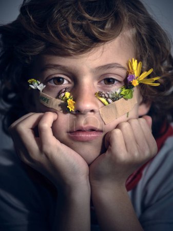 Foto de Foto de niño adorable con flores de colores bajo los ojos unidos a la cara con yesos médicos mirando a la cámara mientras se apoya en las manos - Imagen libre de derechos