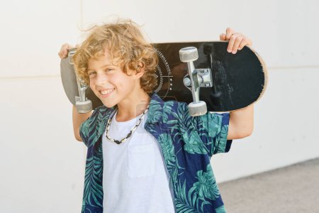 Positive blond kid in casual clothes looking at camera and holding skateboard while standing against street wall on sunny day