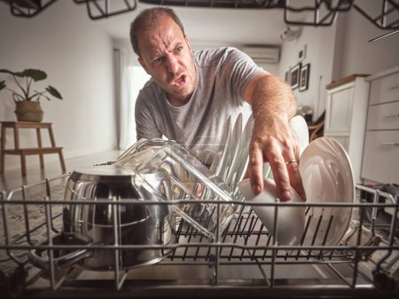 Téléchargez les photos : Furieux mâle en tenue décontractée déchargeant lave-vaisselle moderne avec divers vaisselle propre tout en faisant la routine des travaux ménagers dans la cuisine légère à la maison - en image libre de droit