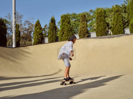 Photo for Side view of boy skater in casual clothes and with sneakers riding skateboard on asphalt road in sunny day in park with green trees background - Royalty Free Image