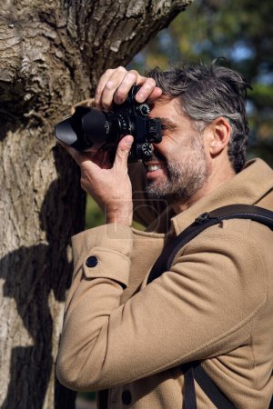 Foto de Vista lateral del fotógrafo masculino con mochila tomando fotos en cámara fotográfica profesional mientras explora el bosque verde en tiempo soleado - Imagen libre de derechos