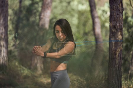 Téléchargez les photos : Femme hispanique concentrée en tenue de sport tirant la bande de résistance tout en faisant de l'exercice pour garder le corps fort dans les bois - en image libre de droit