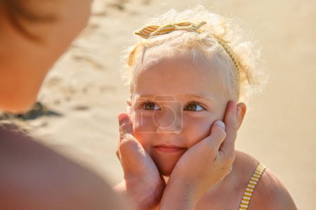 Téléchargez les photos : Angle élevé de mignonne petite fille aux cheveux blonds regardant la mère toucher le visage tendrement sur la plage de sable fin le jour ensoleillé - en image libre de droit