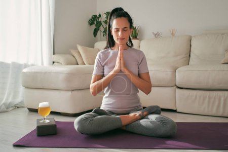 Photo for Full body of peaceful Hispanic female in sportswear meditating with closed eyes in Lotus pose with prayer hands on mat with beer - Royalty Free Image