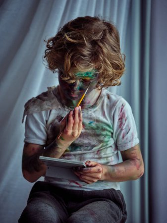 Foto de Niño creativo vistiendo camiseta sucia con manchas de pintura sosteniendo el pincel y pintándose a sí mismo usando el espejo - Imagen libre de derechos