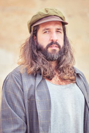 Foto de Retrato de hombre adulto barbudo serio con pelo largo ondulado y bigote en atuendo y gorra inusual - Imagen libre de derechos