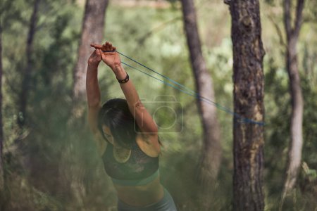 Téléchargez les photos : Femme saine levant les mains au-dessus de la tête tout en tirant la bande de résistance attachée sur le tronc d'arbre pendant l'entraînement actif dans les bois - en image libre de droit