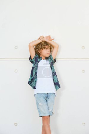 Foto de Niño de moda de confianza en ropa casual elegante mirando hacia otro lado mientras está de pie contra la pared blanca en la ciudad - Imagen libre de derechos