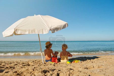 Foto de Vista trasera de niños pequeños irreconocibles sentados en la playa de arena cerca del mar ondulante en el soleado día de verano - Imagen libre de derechos