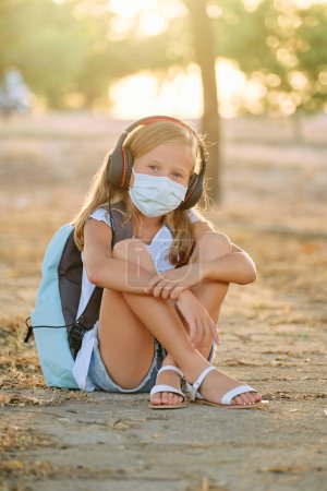 Photo for Full body of child in protective mask sitting on ground and embracing knees while listening to music using headphones and looking at camera in woods during COVID 19 pandemic - Royalty Free Image