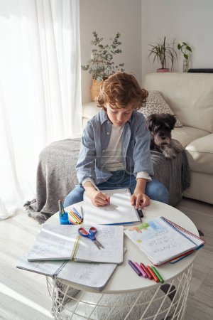 Téléchargez les photos : Petit garçon en vêtements décontractés assis à table et dessin avec crayon près du chien dans le salon pendant le week-end jour à la maison - en image libre de droit