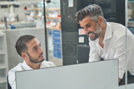 Photo for Content bearded male engineers in formal outfits chatting while browsing modern computer a table during work on project in contemporary office - Royalty Free Image