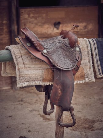Photo for Brown leather western saddle with iron stirrups on metal railing with pads against wooden shabby barn in rural area on summer day - Royalty Free Image