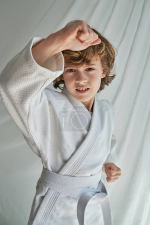 Foto de Niño preadolescente de pelo rizado en kimono blanco de pie cerca de la cortina en posición de defensa mientras practica la técnica de karate y mira a la cámara - Imagen libre de derechos