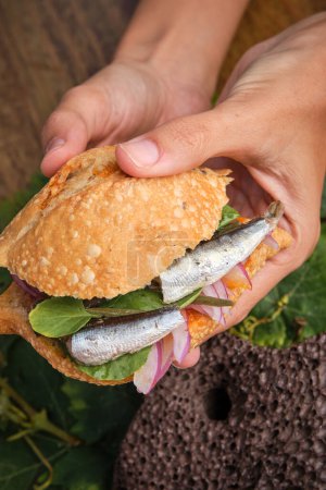 Photo for Vertical photo of a hands grabbing a traditional harvest sandwich with sardines, meat and different vegetables. Gastronomy of Canarias Islands - Royalty Free Image