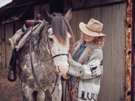 Photo for Female equestrian in cowboy hat and stallion in bridle and western saddle near wooden stable - Royalty Free Image