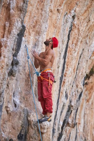 Foto de Vista lateral de cuerpo completo de un fuerte escalador masculino sin camisa con arnés de seguridad frotándose las manos mientras está de pie en la ladera rocosa de la montaña - Imagen libre de derechos