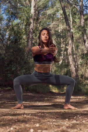 Photo for Full body of barefoot sportive Hispanic female in sportswear outstretching arms forward while doing plie squats during fitness training in woods - Royalty Free Image