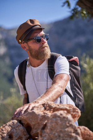Téléchargez les photos : Voyageur masculin sérieux en vêtements décontractés avec sac à dos debout près d'un tas de roches et admirant la nature pendant les vacances d'été - en image libre de droit