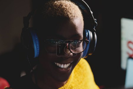 Téléchargez les photos : Portrait de femme afro-américaine joyeuse dans des lunettes aux cheveux courts blancs écoutant de la musique dans des écouteurs et regardant la caméra - en image libre de droit