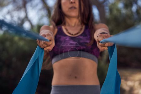 Téléchargez les photos : Cultivez une jeune femelle anonyme aux longs cheveux foncés en vêtements de sport tirant des rubans bleus tout en pratiquant le yoga aérien dans le parc - en image libre de droit
