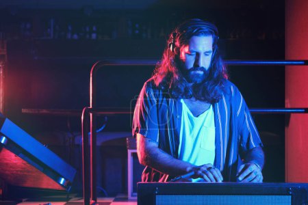 Foto de DJ masculino cualificado serio con barba en ropa casual usando mezclador moderno mientras se realiza la canción en el club nocturno con iluminación tenue - Imagen libre de derechos
