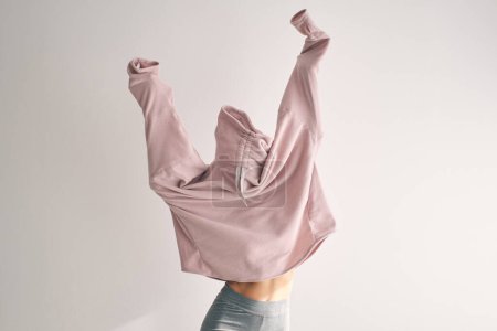 Foto de Cara oculta femenina irreconocible con una elegante prenda holgada mientras levanta las manos contra un fondo blanco - Imagen libre de derechos