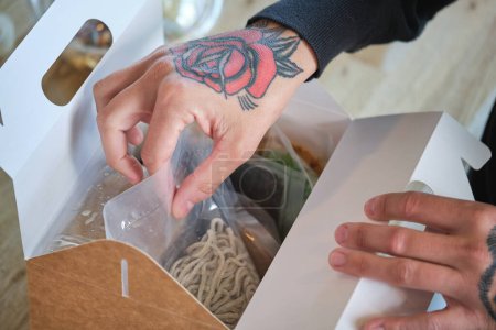 Photo for From above of crop anonymous chef putting raw pasta in plastic wrap into carton box with various ingredients of ramen - Royalty Free Image