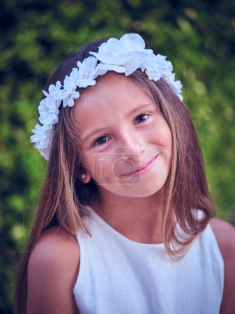 Téléchargez les photos : Charmante jeune fille en robe blanche et couronne florale regardant la caméra tout en se tenant debout sur un fond flou - en image libre de droit