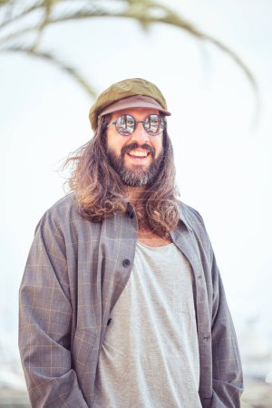 Foto de Hombre barbudo feliz con pelo largo en gorra de moda de pie en la calle soleada con árbol en la ciudad en el día de verano - Imagen libre de derechos