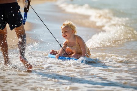 Photo for Full body of smiling girl in yellow swimsuit and headband sitting on blue surfboard and playing while someone dragging her among splashing waves on sandy seashore - Royalty Free Image