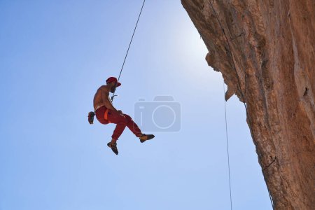 Photo for Full body side view of strong shirtless male alpinist hanging on rope while descending from rocky cliff against blue sky - Royalty Free Image