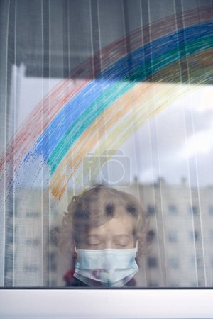 Foto de A través de la vista de cristal del niño en la máscara protectora de pie solo con los ojos cerrados cerca de la ventana con cortinas durante la pandemia de coronavirus - Imagen libre de derechos