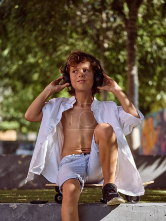Zufriedener Junge sitzt auf Skateboard und schaut weg, während er Kopfhörer berührt und im Sommer im grünen Park Musik hört