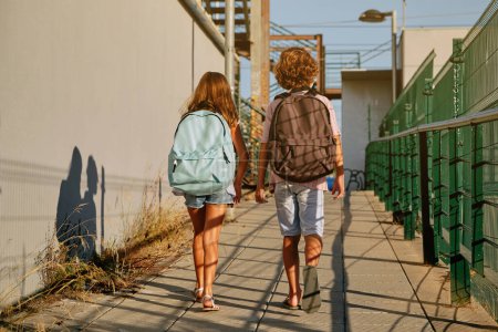 Foto de Vista trasera de niño y niña irreconocibles en ropa casual con mochilas caminando a lo largo del camino de asfalto juntos en la calle de la ciudad - Imagen libre de derechos