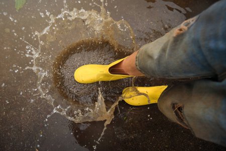 Foto de Vista superior de una persona irreconocible en botas de goma amarillas salpicando agua en un charco en un día lluvioso de otoño en la naturaleza - Imagen libre de derechos