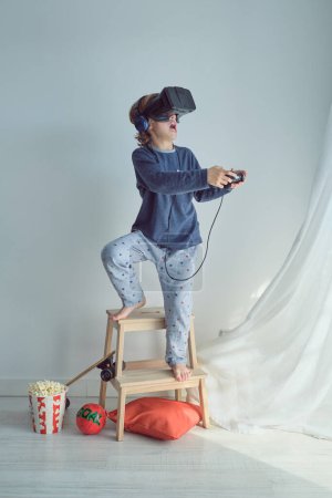 Foto de Cuerpo completo de preadolescente descalzo en ropa de dormir y VR auriculares y auriculares jugando videojuego con joystick de pie en la escalera de taburete - Imagen libre de derechos