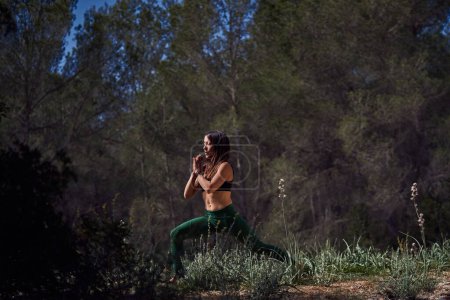 Foto de Mujer hispana pacífica practicando Crescent Lunge y manteniendo las manos juntas en el pecho mientras respira con los ojos cerrados en el paisaje rural - Imagen libre de derechos